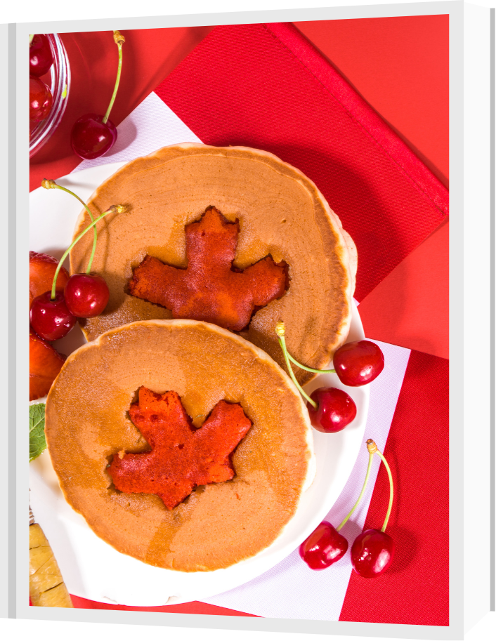Picture of pancakes with a maple leaf cut in the centre of them, with berries around them. The pancakes and berries are on top of a Canadian Flag tablecloth